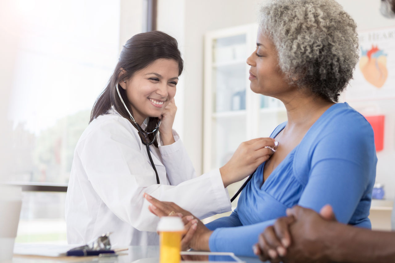 A smiling female doctor sits at a table next to a senior female patient. She uses her stethoscope to listen to her patient's lungs. There is a clipboard and prescription bottle on the table. The hands of another person can be seen resting on the table in the foreground.