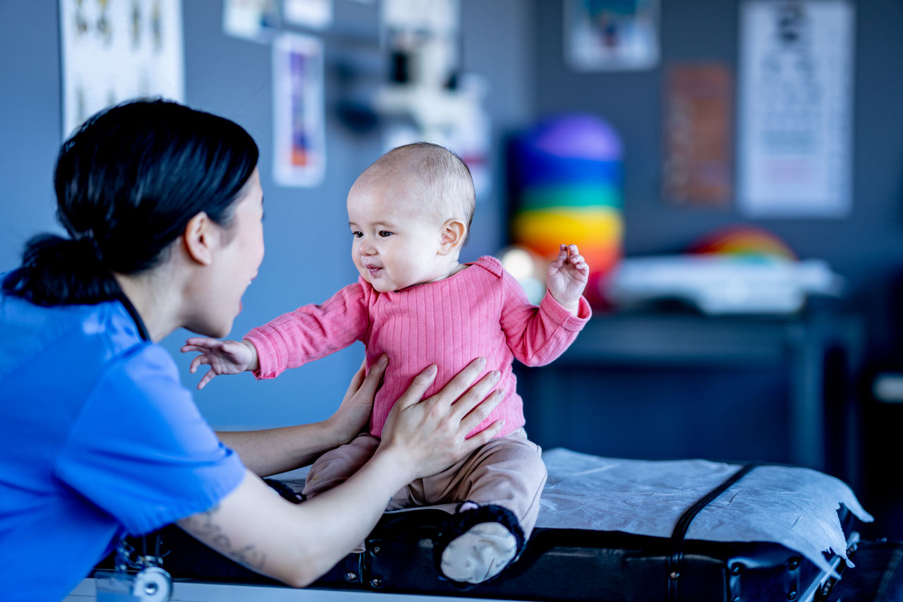 A sweet little baby girl sits up on a medical exam table with the assistance of a nurse during a routine well-baby check-up.  She is dressed casually and has a smile on her face as her female nurse, of Asian decent, checks her over.