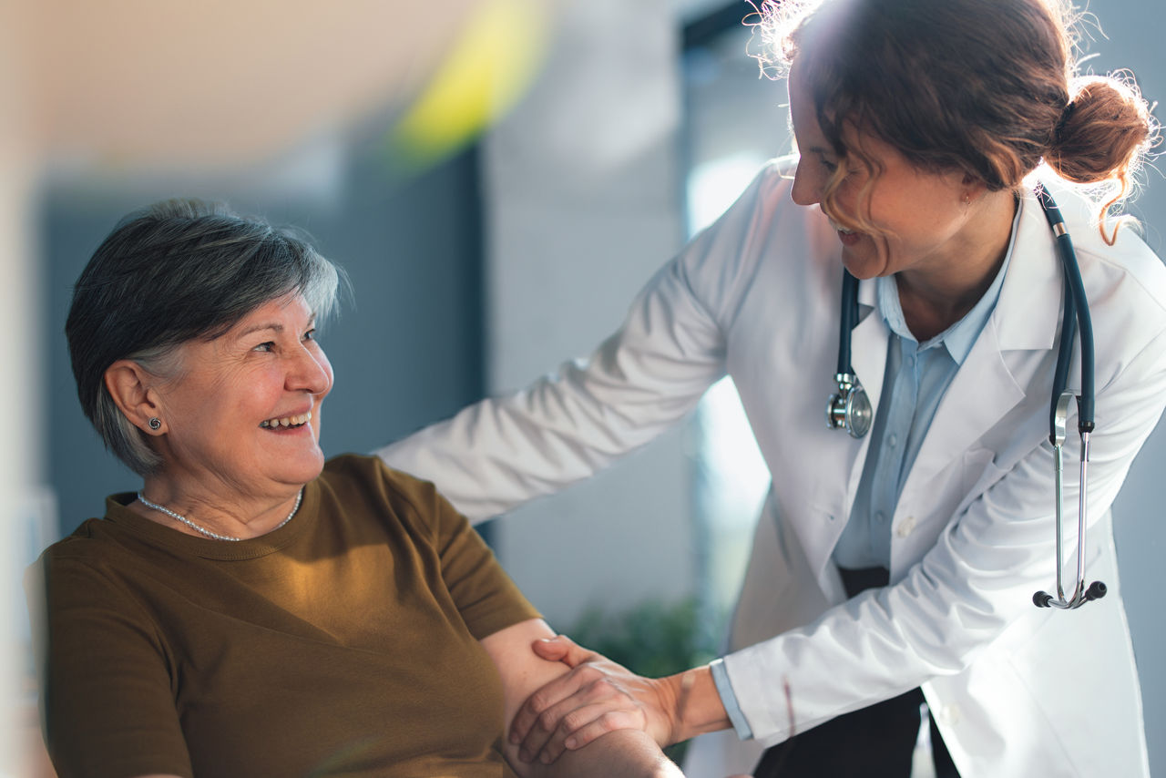 Senior woman smiles while a friendly female doctor in a white coat comforts her in a bright doctor office, conveying trust, compassionate medical care and positive patientâprovider connection.