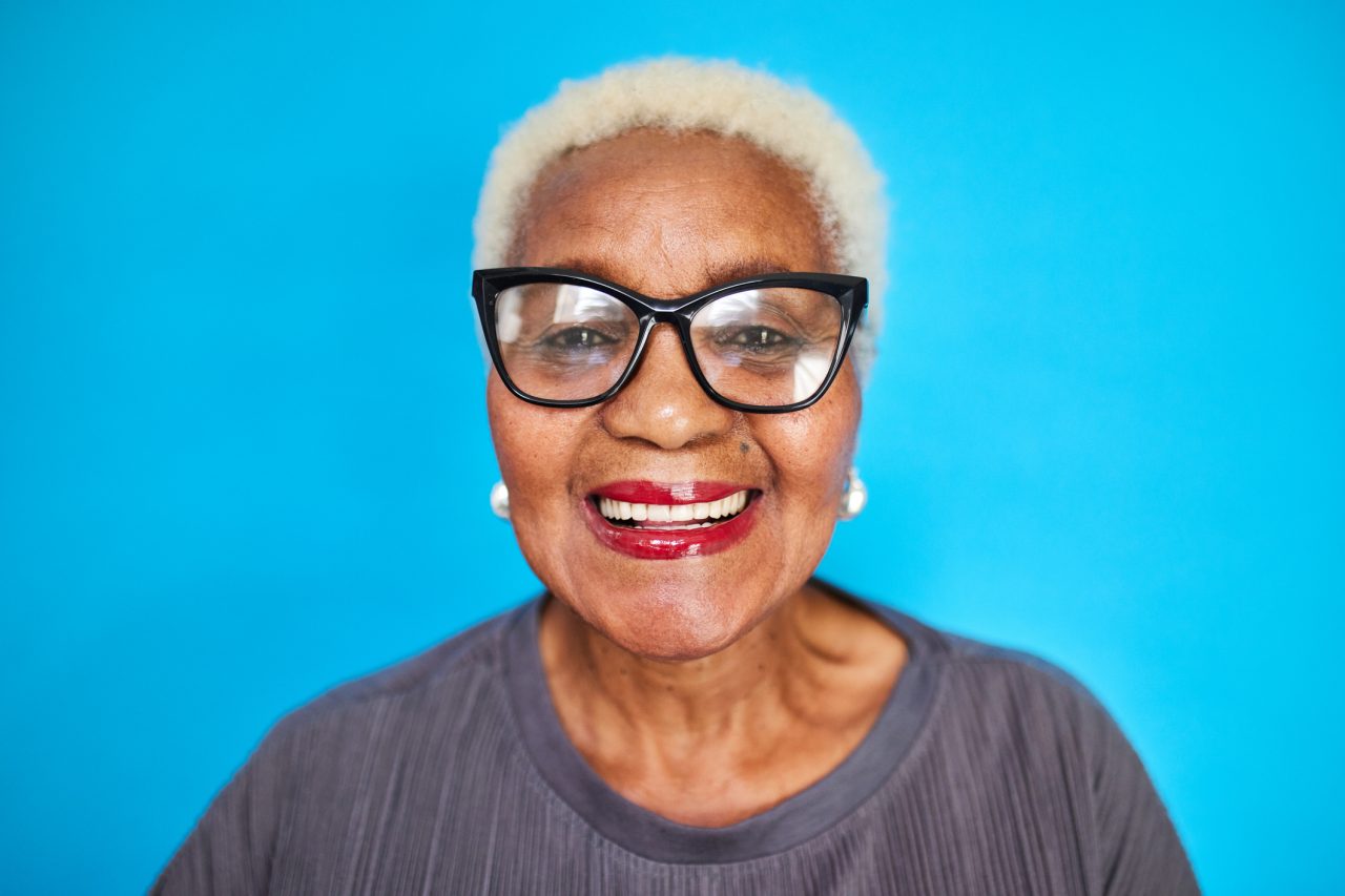 Close-up of a cheerful senior woman with glasses, bright red lipstick, and white hair against a vibrant blue backdrop, radiating joy and warmth.